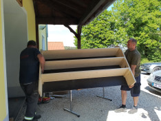 Két férfi cipel egy 2,5*1,5 méteres fábol készült cipőtartót.